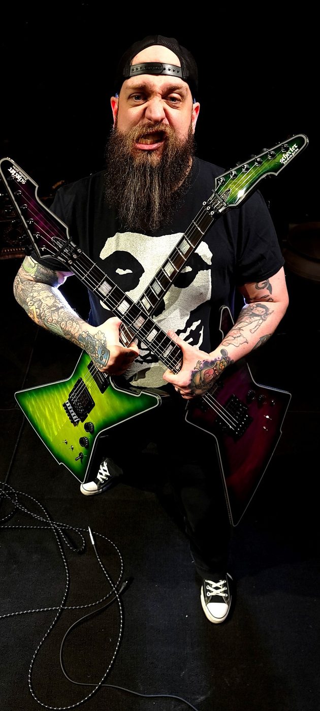 Nate Laff snarling while holding two crossed Schecter Explorer-style guitars, one green and one purple